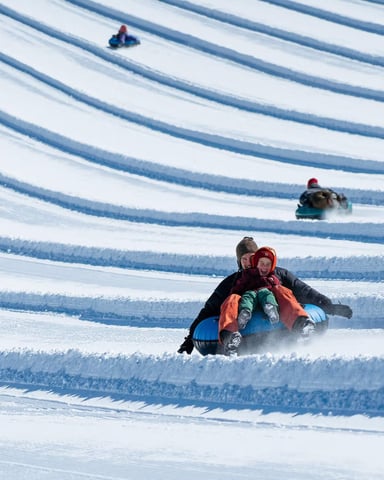 Multiple people sledding down snowy hill with parallel grooved tracks