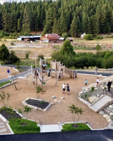 Aerial view of large playground with climbing structures, swings, and sandbox beside river, with ranch buildings and evergreen forest beyond