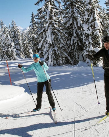 Two cross-country skiers on groomed trail through snow-covered evergreen forest with snow-capped mountain peaks in background