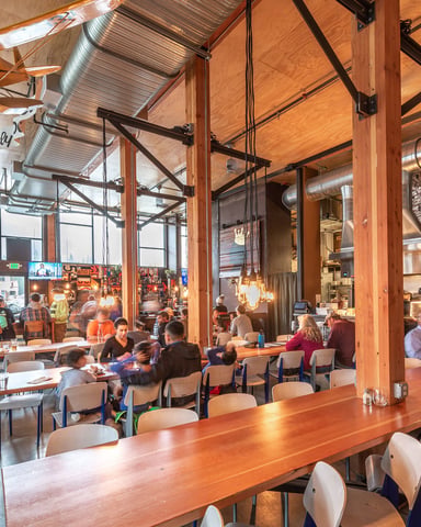 Industrial restaurant with exposed wood beams, open kitchen, communal wooden tables, and diners seated throughout the space