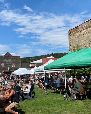 Outdoor farmers market event with crowds under white and green tents, historic stone building and wooden structures in background