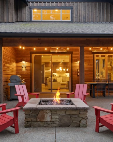 Modern house exterior with stone fire pit, red Adirondack chairs, and illuminated covered patio at dusk