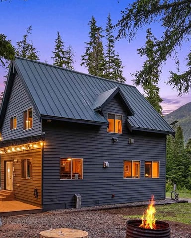 Modern dark cabin with metal roof and lit windows surrounded by tall evergreen trees and mountains at dusk, fire pit in foreground.