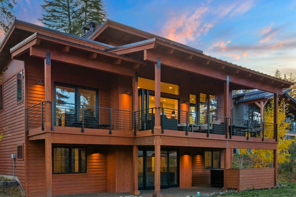 Modern two-story wood cabin with metal railings, large windows, and covered deck surrounded by evergreen trees at dusk.