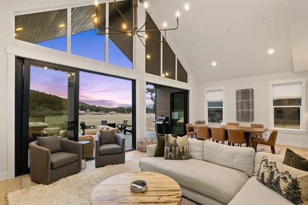 Modern open-plan living room with high ceilings, large windows overlooking meadow and forest, gray sectional sofa, leather armchairs, and dining table.