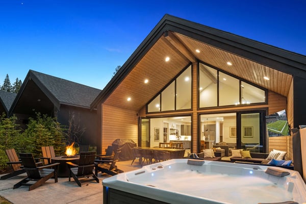 Modern home exterior at dusk with hot tub, fire pit, Adirondack chairs, and illuminated interior visible through large windows