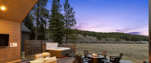 Modern wood cabin patio at dusk with hot tub, fire pit, seating, and forest meadow views at twilight