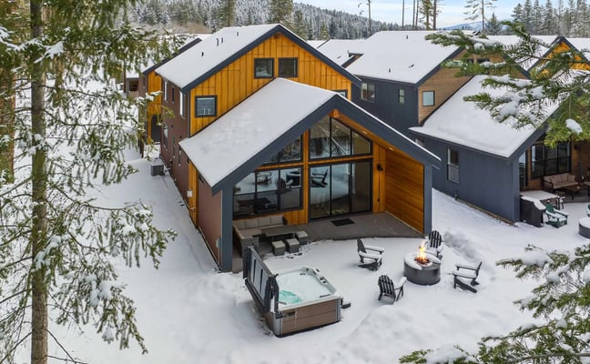 Modern mountain cabins with wood and dark siding in snowy forest clearing, hot tub and fire pit in foreground.