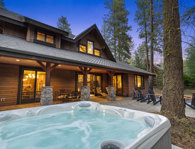 Modern cabin with hot tub, stone patio, and tall pine trees at dusk with lit windows